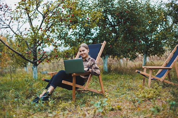 Stijlvolle vrouw zittend op een stoel in de buurt van groene planten in de tuin en op internet surfen op een laptop terwijl ze naar de zijkant kijkt Werk op afstand vrouwelijke freelancer