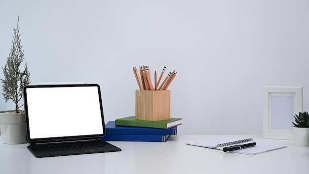 Stijlvolle werkplek met computertablet, plant, notitieboekje, potlodenhouder en boek op wit bureau.