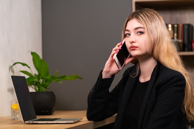 Stijlvolle zakenvrouw aan het chatten op een mobiele telefoon in een kantoor zittend aan haar lamptop en zich met een serieuze uitdrukking op de camera werpend