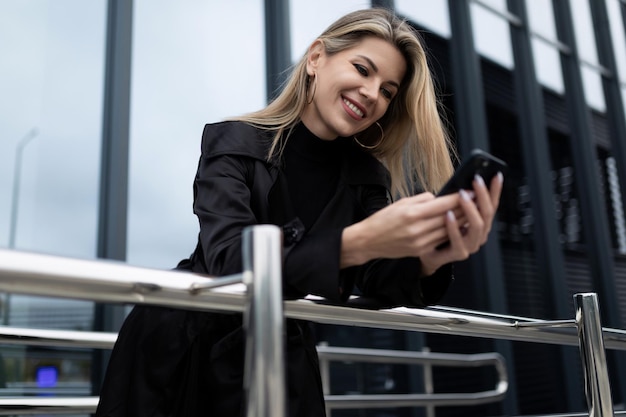 Stijlvolle zakenvrouw op de achtergrond van een glazen gevel met een telefoon in haar handen glimlachend