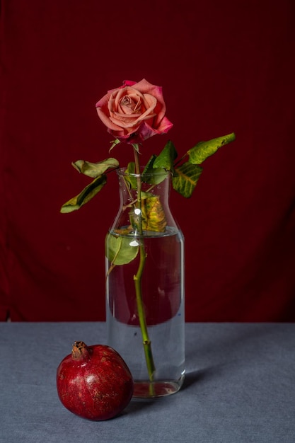 Photo still life on a blue background with red,  red rose in a glass vase and pomegranate,