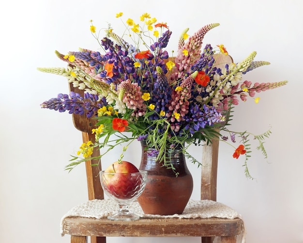 Still life bouquet with lupine and buttercups in the jug and apples in a vase on the chair on a light background.