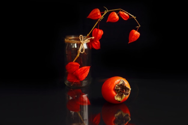 Still life a branch of physalis in a glass jar next to an orange persimmon on a dark background reflection from objects