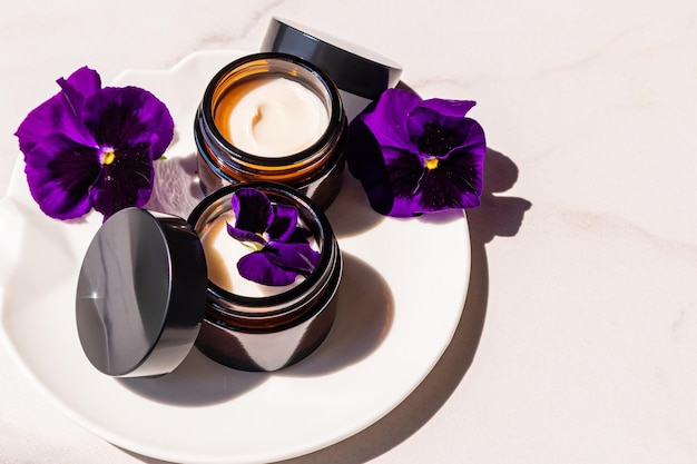 Still life cosmetic Natural cream for facial skin care in open cosmetic jars made of dark glass rejuvenating effect marble background violets
