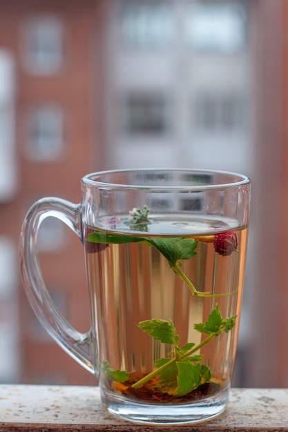 Still life cup with herbal tea. Red house with a lot of windows on the background. Strawberry leaves and berries are floating in the cup. Side view. Vertical frame.