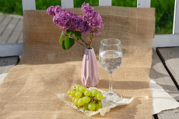 Still life and food photo Bunch of grapes glass of water and vase with lilacs stand on burlap