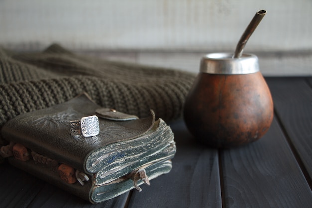 Still life of Hand Crafted Artisanal Yerba Mate Tea Leather Calabash Gourd with Straw, leather notebook, sweater and ring on a black painted table, 