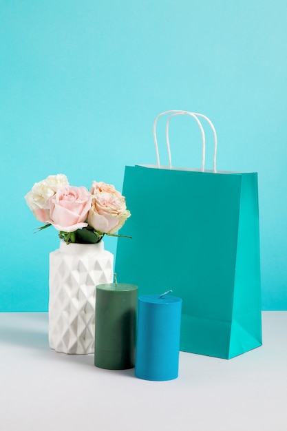 花瓶、キャンドル、紙袋に花のある静物画像。クラフトショッピングバッグのモックアップ。販売や割引のコンセプトです。ブランディングのモックアップ。青色の背景に装飾ショップのコピースペースの画像