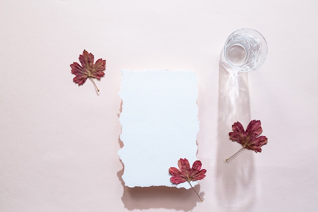 Scena di natura morta con un foglio di carta bianco e foglie colorate autunnali e un bicchiere d'acqua di cristallo