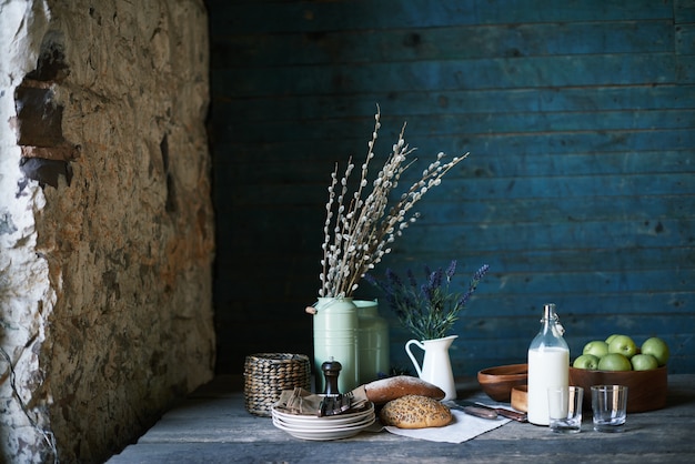 Still life of table served for breakfast with homemade bread, fresh milk in jug and apples in wooden bowl, stack of plates, glasses and napkins