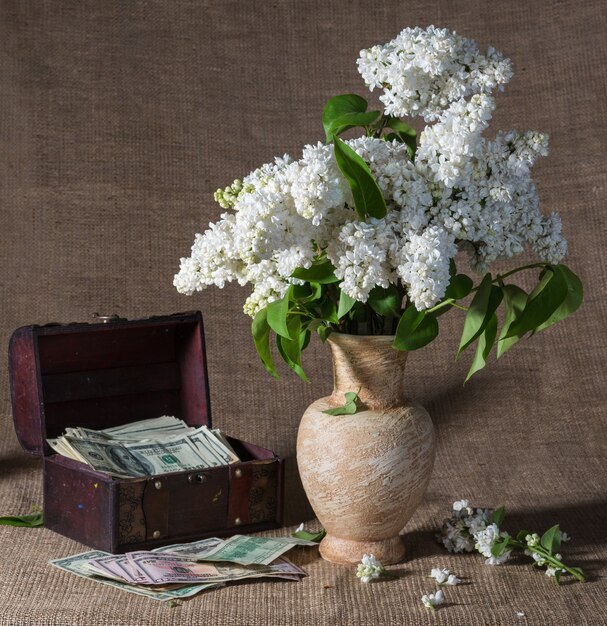 Still life with  blooming branches of lilac in vase and dollars in chest