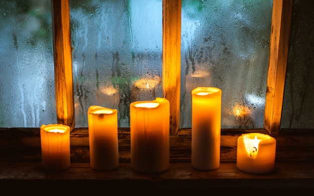 Still life with burning candles in an old country house near a wooden wet window in the autumn evening.