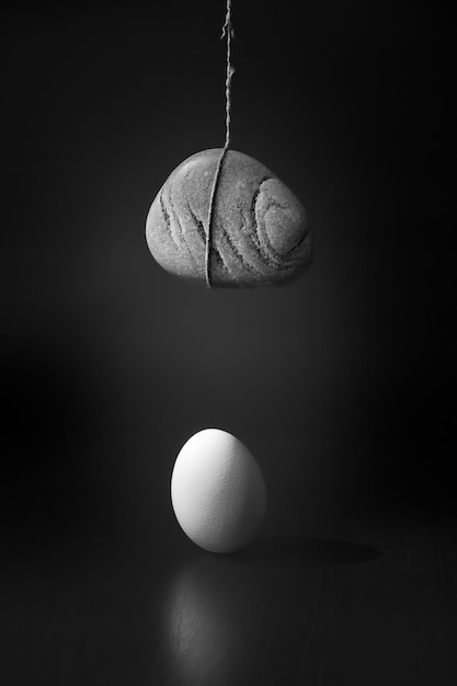 Photo still life with egg standing on wooden table and cobblestone hanging over it on thin tearing thread