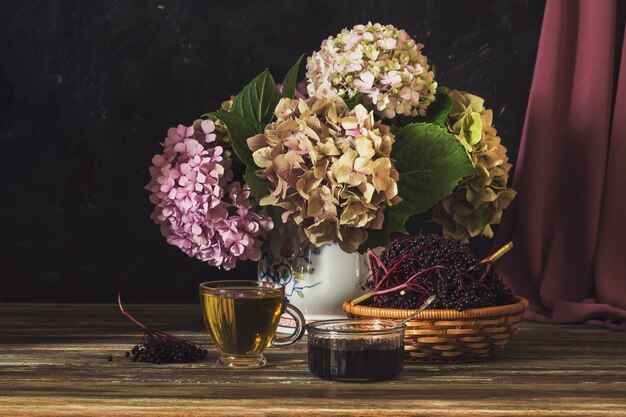 Still life with gentle hydrangea a cup of tea and elderberry jam and a basket of elderberries on a wooden table