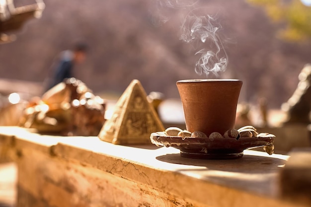 写真 裸の山と砂漠の背景に粘土のカップで熱い東洋のコーヒーを飲む静物 屋外のロードカフェのピラミッド形の記念品の隣のバーにコーヒーカップが座っています