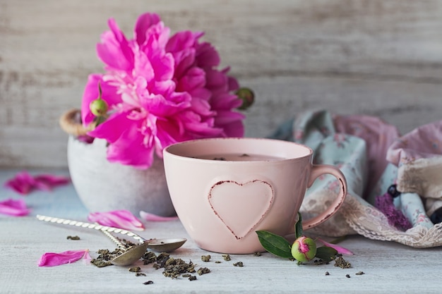 Still life with pink peony flowers and a cup of herbal or green tea on rustic wooden background