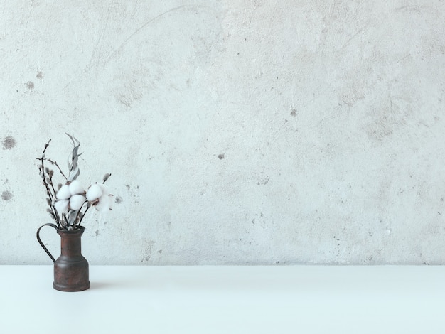 Natura morta con una piccola brocca di rame con un mazzo di fiori secchi su un tavolo bianco con un muro di cemento. fiori di cotone, eucalipto, salice.