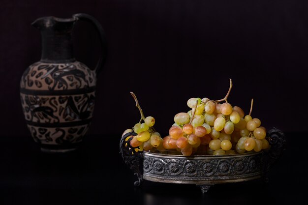 Still life with white grapes and antique pitcher on black background.