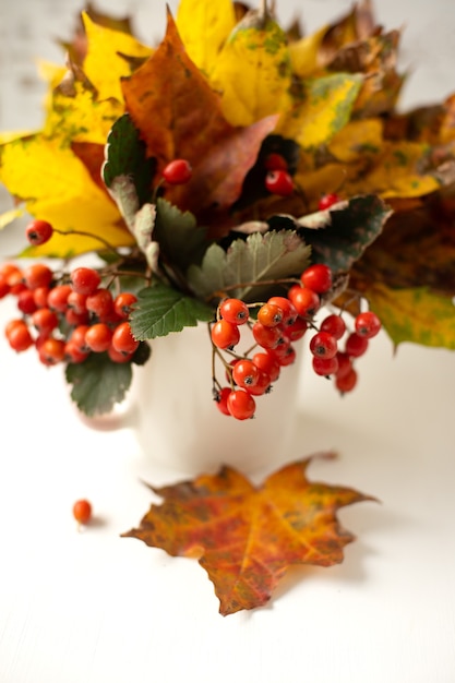 Stilleven. Herfstboeket van heldere gevallen bladeren en rode lijsterbes in een witte kop op een houten achtergrond. Verticale foto