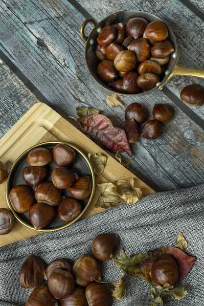 Foto stilleven met metalen braadpan vol kastanjes en gedroogde herfstbladeren op grijs plankoppervlak en bamboetafel met theedoek