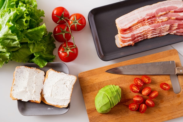 Stillevensamenstelling bestaande uit twee sandwiches, verse tomaten, sla, avocado en dienblad met plakjes spek op keukentafel