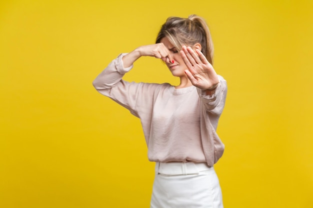 Stinky smell Portrait of young dissatisfied woman with fair hair in casual beige blouse standing pinching her nose with disgust and gesturing stop indoor studio shot isolated on yellow background