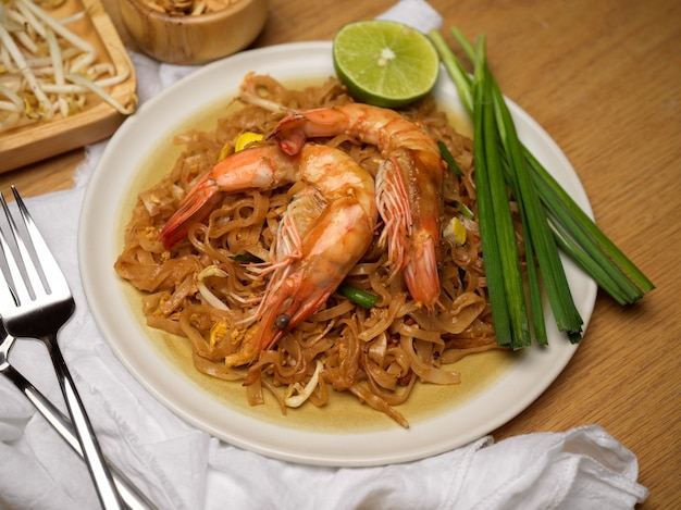 Stir fired Thai noodles with shrimps serving on wooden dinning table with lime, beansprouts and chives, Thai traditional food