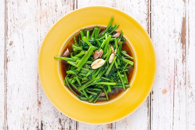 Stir fried water spinach with garlic in yellow ceramic plate on white old wood texture background, top view, light and airy food photography, morning glory