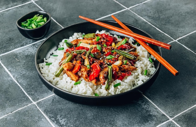 Stir fry with chicken meat vegetables and rice in black bowl on dark tiled stone background