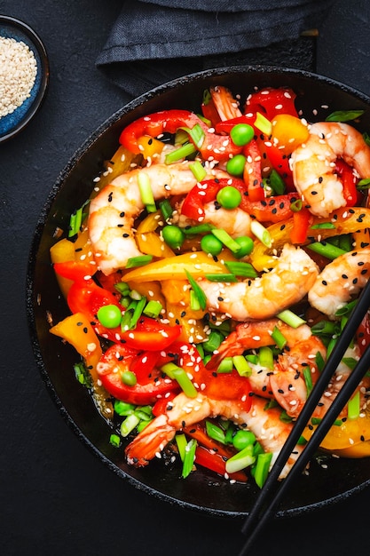 Stir fry with shrimps red and yellow paprika green pea chives and sesame seeds in frying pan Asian cuisine dish Black stone kitchen table background top view