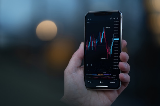 Stock exchange and trading online. Male hand holding smartphone and using investment app, analyzing market data in real time. Selective focus on mobile phone with financial graph chart on screen