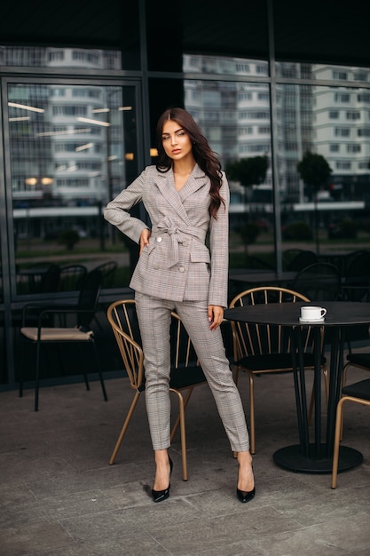 Foto stock foto van een trendy model in geruit beige pak en zwarte hakken met lang golvend bruin haar poseren naast zwarte tafel en stoel buiten restaurant of café