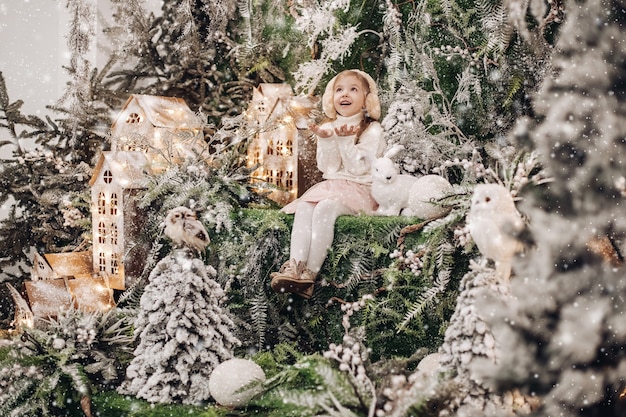 花輪で照らされた手作りの家とクリスマスの森に囲まれた横に白いおもちゃのウサギと降雪の下に座っているイヤーマフの美しい少女のストックフォト。