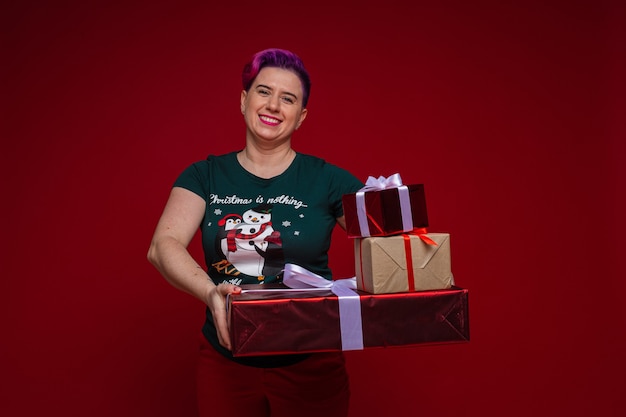 Stock photo of cheerful adult woman with short dyed hair wearing Christmas t-shirt, holding stack of beautiful wrapped Christmas gifts in hands and smiling