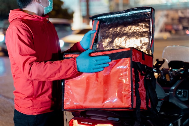 Stock photo of a food deliveryman in red uniform carrying a food delivery box to deliver for customer for order during COVID-19 pandemic and  lockdown in the city at night time in Thailand.