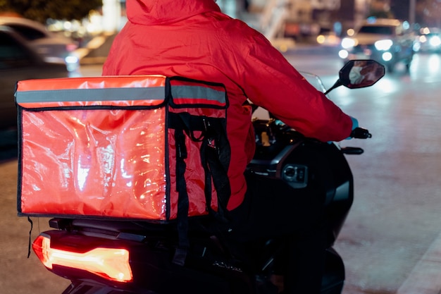 COVID-19パンデミックとタイの夜間の都市での封鎖の間に注文のために顧客に配達するために食品配達ボックスを運ぶ赤い制服を着た食品配達人のストックフォト。