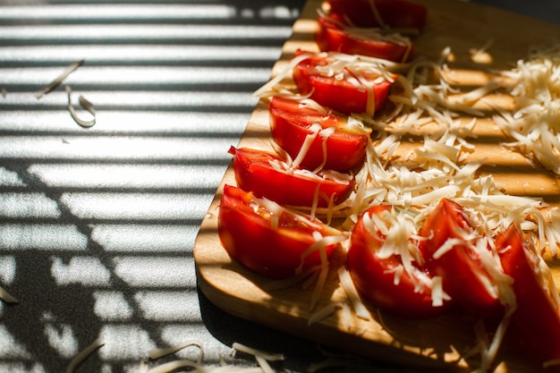 写真 日光の下で木の板に細かく刻んだチーズとスライスしたトマトのクローズアップのストックフォト。テーブルの上のブラインドシャドウ。調理プロセス。