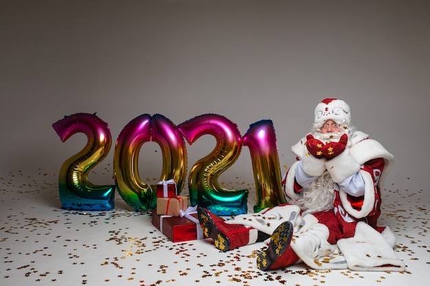 Foto di stock di jolly father frost in abiti festosi con una lunga barba bianca che allunga le braccia e sorride alla telecamera sotto i coriandoli dorati volanti. isolato su sfondo grigio.