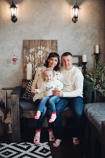 Stock photo portrait of cheerful Caucasian family with little daughter on knees snuggling sitting on small couch under snowfall in cozy room. They are looking at camera, embracing their daughter.