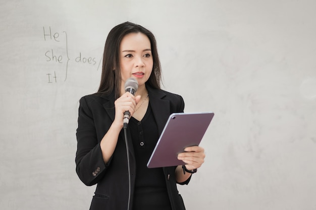 Foto di stock ritratto di un'insegnante donna asiatica allegra fiduciosa in un'uniforme nera con un tablet digitale e un laptop per insegnare la lingua moderna in classe