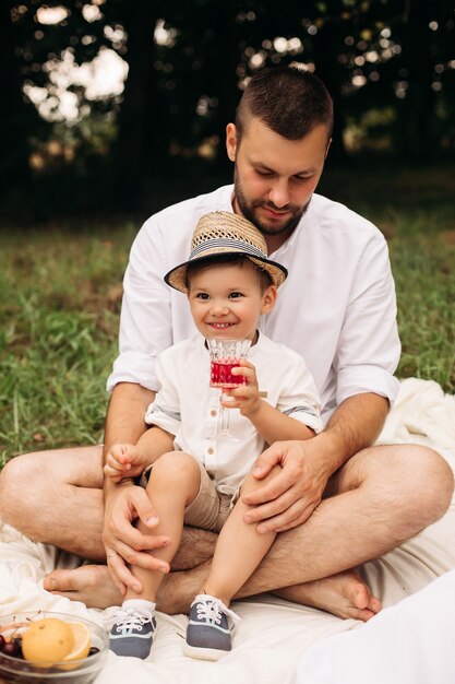 Stock photo portrait of a happy little boy in summer hat drinking juice from a glass sitting with his bearded father on the blanket at picnic on summer day.