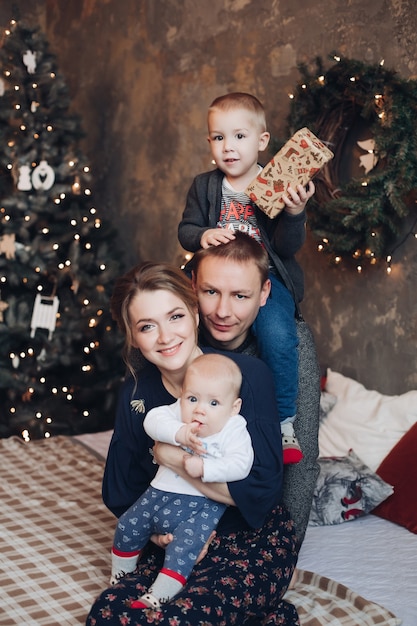 Фото портрет веселой семьи с двумя сыновьями, держащими подарки, сидя на кровати под снегопадом и улыбаясь в камеру