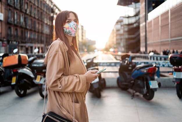 거리에서 그녀의 스마트 폰을 사용하는 젊은 백인 여자의 포토. 그녀는 코로나 19로 인해 안면 마스크를 쓰고 있습니다.