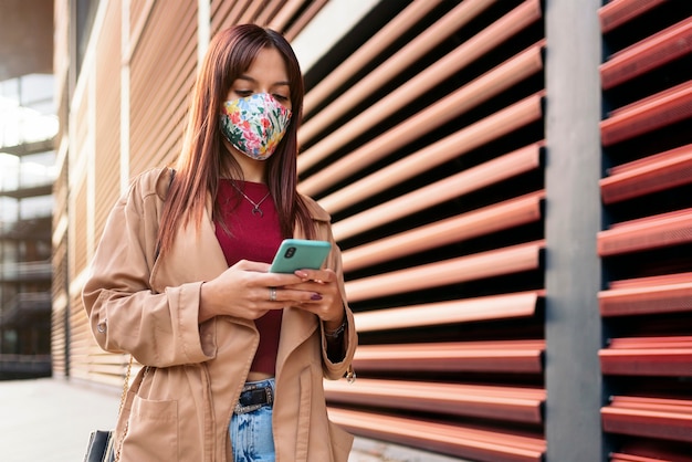 거리에서 그녀의 스마트 폰을 사용하는 젊은 백인 여자의 포토. 그녀는 코로나 19로 인해 안면 마스크를 쓰고 있습니다.