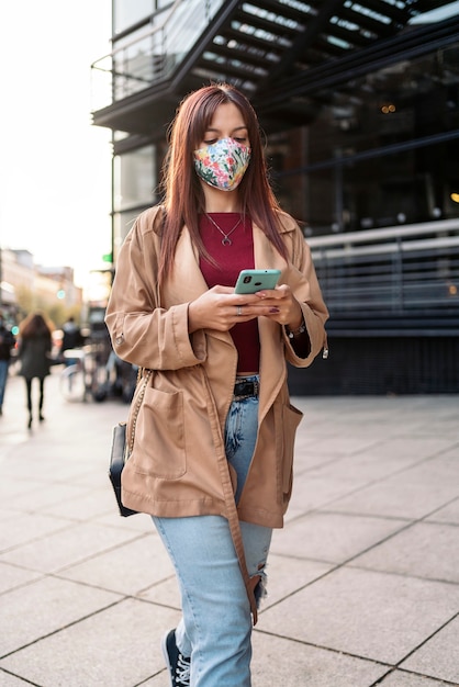 Фондовый фото молодой кавказской женщины, использующей свой смартфон на улице. На ней маска для лица из-за covid19.