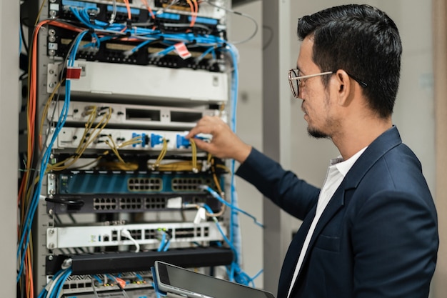 Фондовый фото молодой сетевой техник, держащий планшет, работающий для подключения сетевых кабелей в серверном шкафу в сетевой серверной комнате. ИТ-инженер, работающий в сетевой серверной комнате