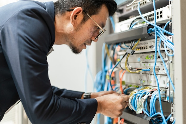 ネットワークサーバールームのサーバーキャビネットでネットワークケーブルを接続するために働いているタブレットを持っている若いネットワーク技術者のストックフォト。ネットワークサーバールームで働くITエンジニア