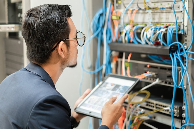 ネットワークサーバールームのサーバーキャビネットでネットワークケーブルを接続するために働いているタブレットを持っている若いネットワーク技術者のストックフォト。ネットワークサーバールームで働くITエンジニア