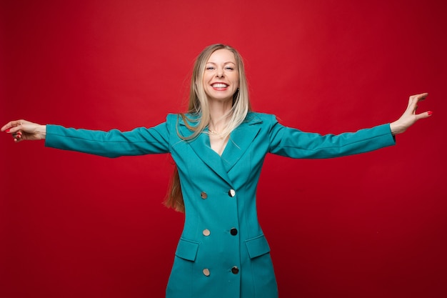 Stockfoto portret van gelukkige joviale blonde vrouw in casual double-breasted jurk of jas met uitgestrekte armen glimlachend in de camera. plezier hebben in de studio op rode achtergrond.