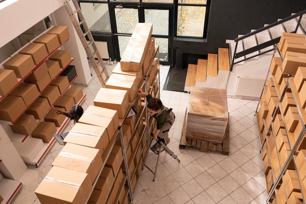 Stockroom worker standing on ladder checking carton boxes, preparing customers orders before start shipping packages in warehouse. Supervisor checking goods inventory in storehouse